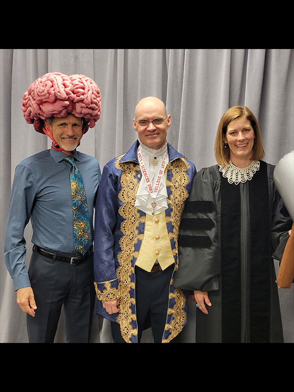 A photo of three people in costume, posing and smiling. On the left, a man wears a large pink brain hat and a blue shirt with a science-themed tie. In the center, a man wears a colonial-style outfit with a gold-embroidered blue coat and a medallion that reads "College of Arts + Sciences 2023." On the right, a woman wears an academic robe.