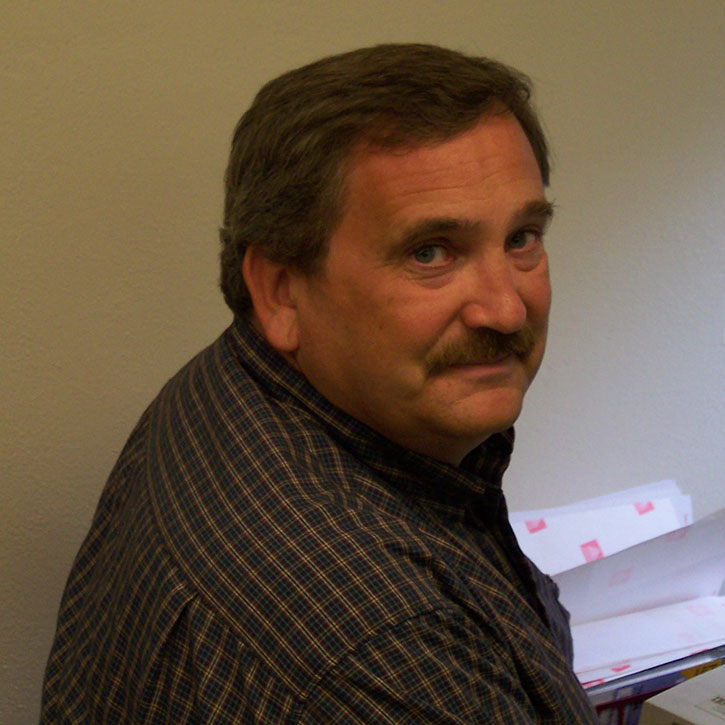A photo of Bill Head, who wears a plaid shirt and sits at a desk with papers. He has light brown hair and a mustache and is turned slightly toward the camera with a gentle smile.