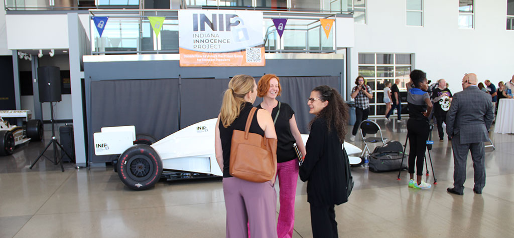 A photo taken at an event for the Indiana Innocence Project. Three women stand in the foreground, smiling and talking, while a white racecar with INIP branding is parked behind them. Other attendees are scattered in the background in a modern, sunlit atrium.