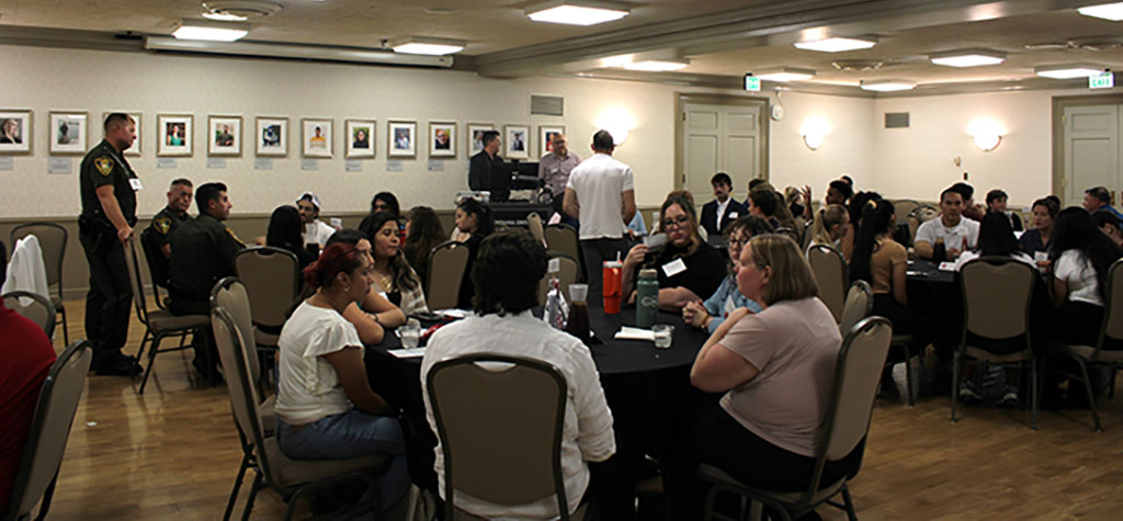 A photo of a large networking event in a banquet hall. Multiple round tables are filled with students, faculty, and uniformed officers. People are eating, talking, and interacting in a warmly lit room with framed photos on the walls.