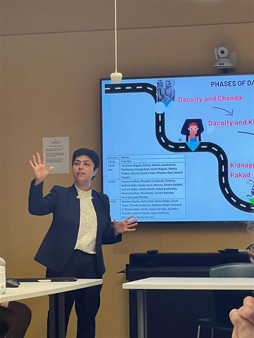 A photo of Deepika Suri giving a presentation in a classroom. She gestures with one hand while standing in front of a screen that displays a diagram and text about crime phases. She wears a navy blazer and white polka-dotted blouse.