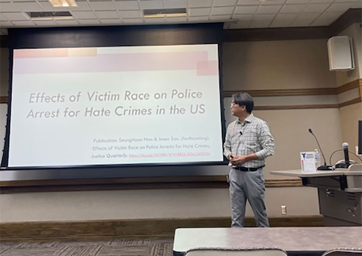 A photo of Simon Han delivering a lecture in a classroom. He stands next to a projection screen displaying the title of his research: “Effects of Victim Race on Police Arrest for Hate Crimes in the US.” He wears glasses and a checkered shirt.