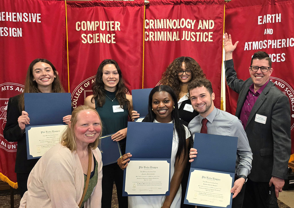 A group photo of students and a faculty member posing with certificates in front of red academic banners labeled “Comprehensive Design,” “Computer Science,” “Criminology and Criminal Justice,” and “Earth and Atmospheric Sciences.” Everyone is smiling proudly.
