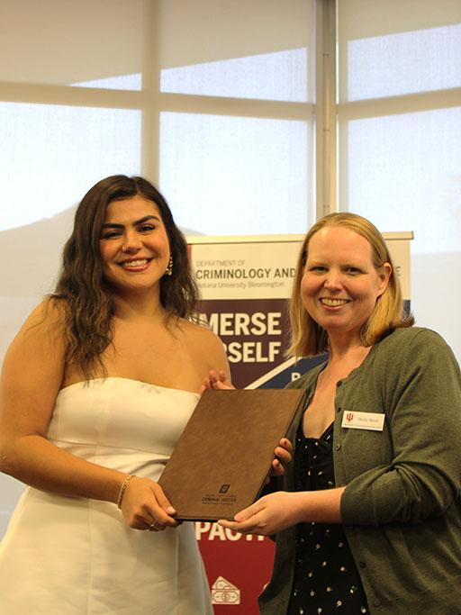 A photo of two women smiling and holding a certificate or folder. One wears a white dress, and the other wears a green cardigan. They stand in front of a banner for the Department of Criminology and Criminal Justice at Indiana University Bloomington.