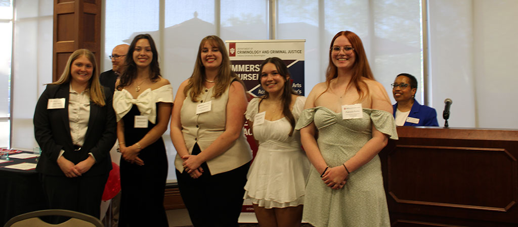 A photo of five students standing in a row, smiling at the camera during a recognition event. They are dressed in semi-formal attire and stand in front of a banner for the Department of Criminology and Criminal Justice. Two staff or faculty members stand at the podium in the background.