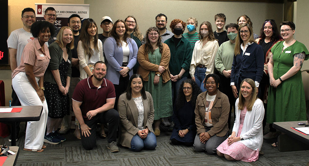 A group photo of about 30 graduate students, faculty, and staff posing in a classroom. Some people are standing, while others are kneeling in front. A departmental banner is visible in the background.