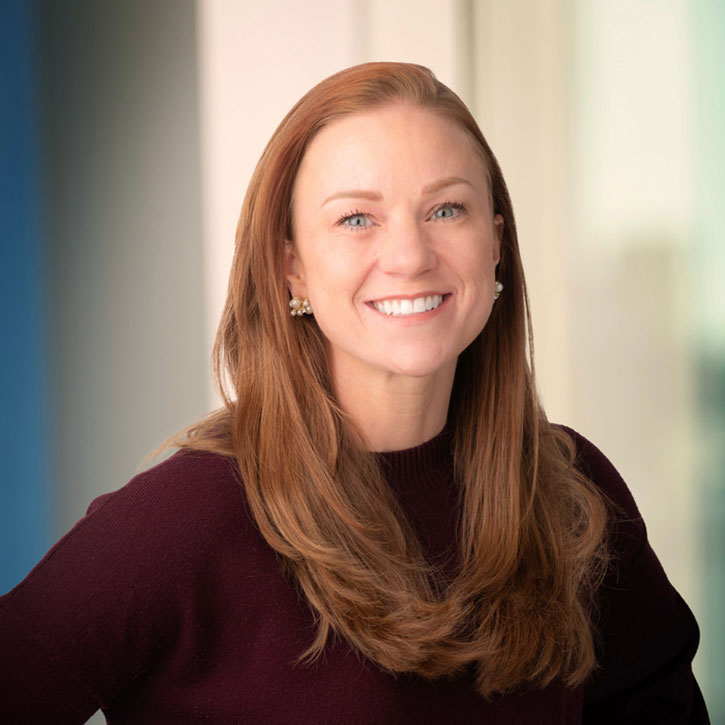 A headshot of Deanna Dwenger, who has long red hair and is smiling in a well-lit indoor space. She wears a maroon top and pearl earrings.