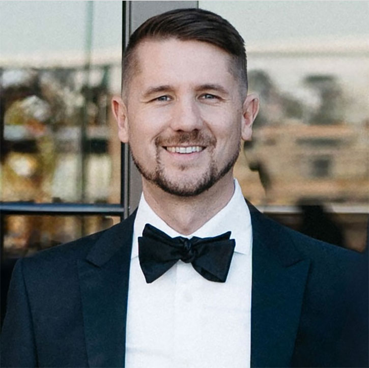 A headshot of Nick Ackerman, who wears a tuxedo with a black bow tie, smiling in front of a glass doorway with reflections of trees and buildings.