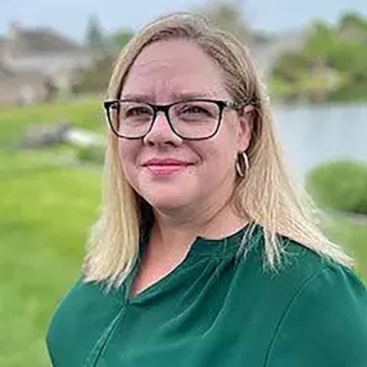 A headshot of Shelley Bizila, who wears glasses and a green blouse, smiling outdoors in front of a grassy area and a body of water.