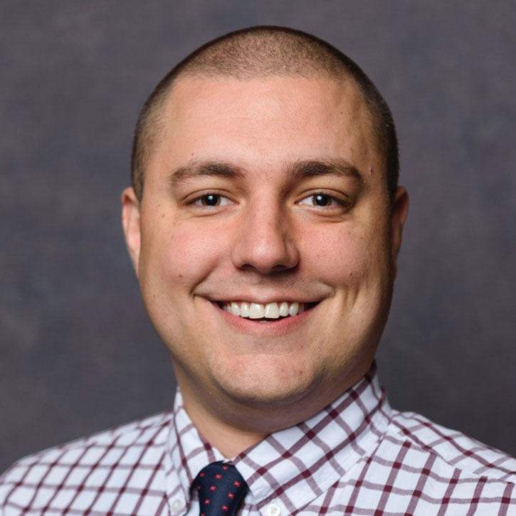 A headshot of Ghazi Tehrani, who wears a checkered shirt and navy tie, smiling against a dark studio-style background.