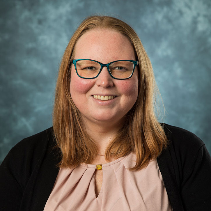 A headshot of Molly Block, who has reddish-blonde hair and teal-framed glasses, smiling in front of a mottled blue-gray studio background.