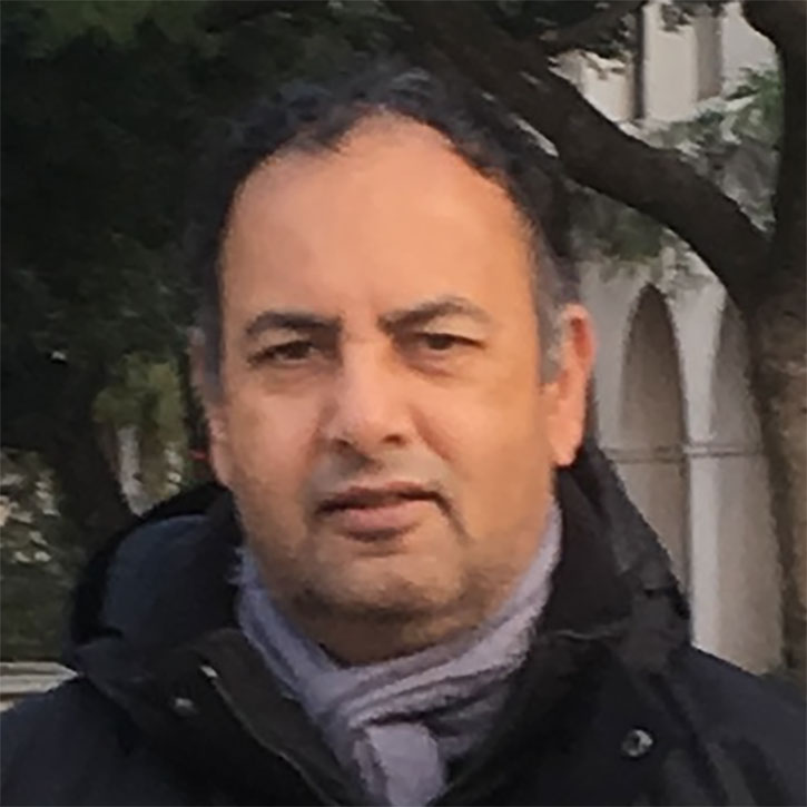 A photo of a man wearing a black jacket and scarf, standing outdoors with blurred buildings and trees in the background.