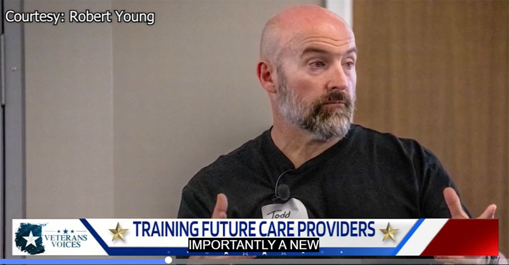A news screenshot of Todd Burkhardt speaking during a televised segment titled “Training Future Care Providers” on Veterans Voices. He is bald with a graying beard and wears a black shirt and microphone headset.