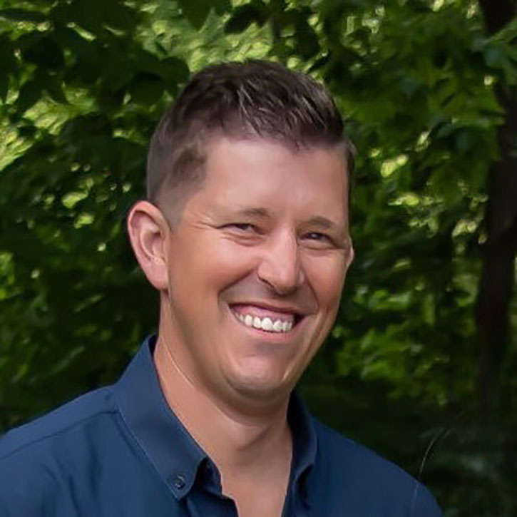 A headshot of Jon Brauer, who poses in a wooded setting and wears a short-sleeved blue button-down shirt.