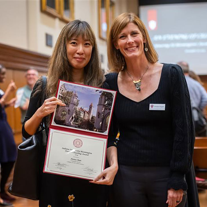 A photo of Fiona Chan holding a red IU award folder with a certificate, standing next to Natalie Hipple. They are in a formal event space with others in the background.