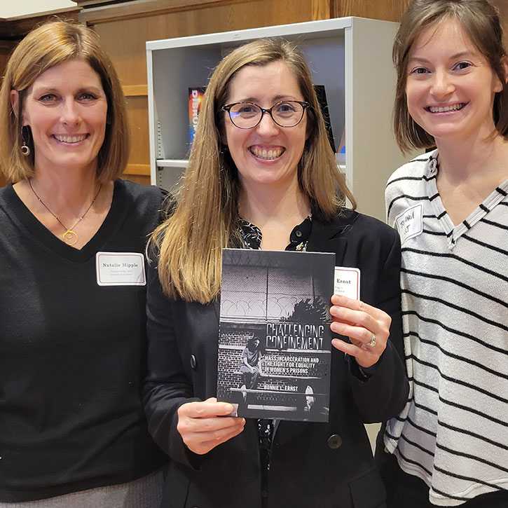 A photo of three women smiling. The woman in the center holds up a book titled “Challenging Confinement.” All are dressed professionally and stand in a room with bookshelves.