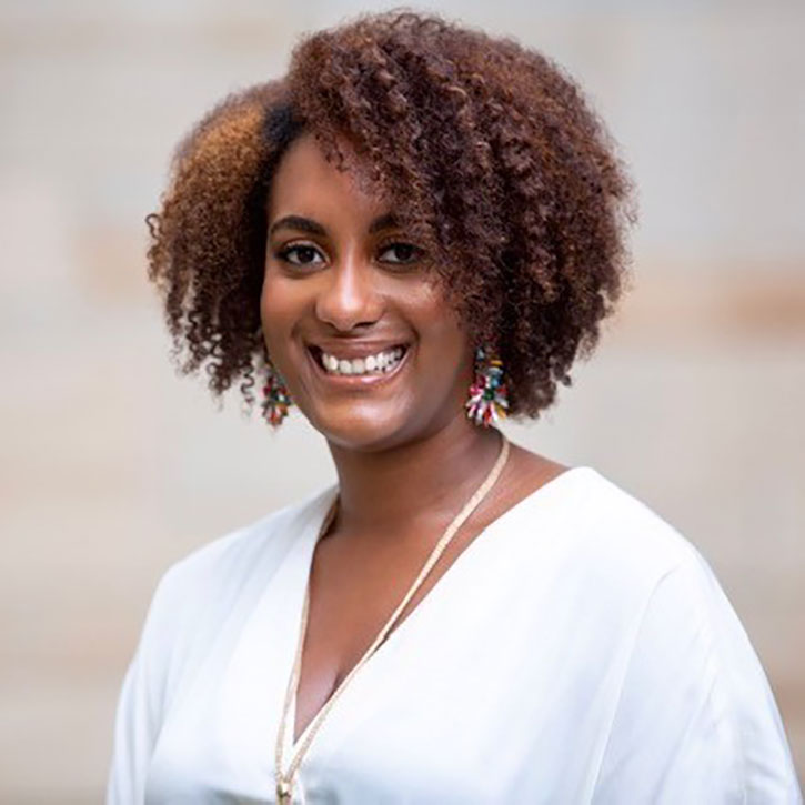 A headshot of a woman smiling in a white blouse with colorful earrings, photographed in soft outdoor light.