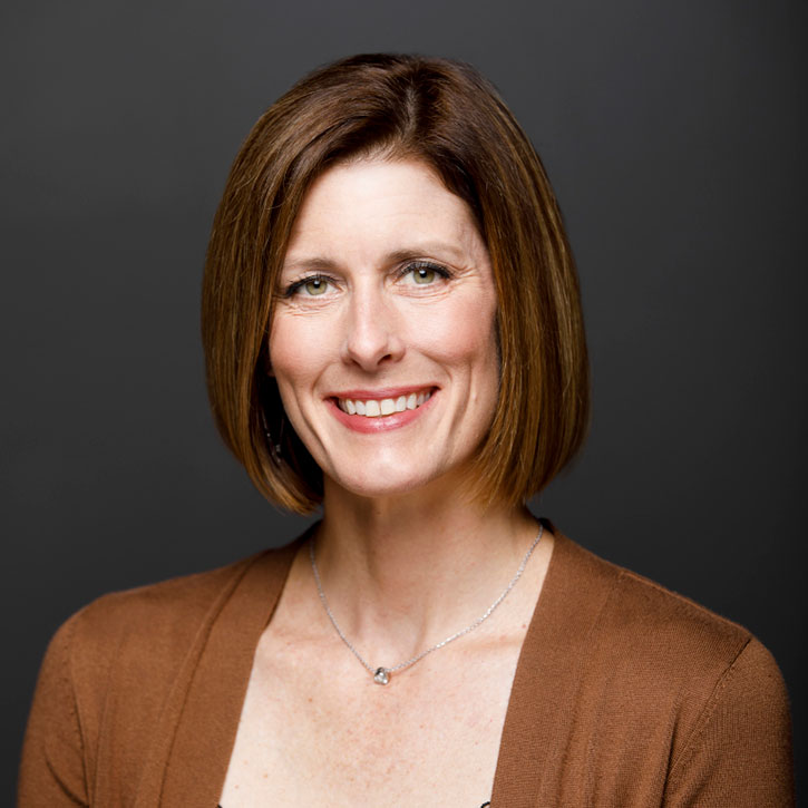 A headshot of a woman with chin-length hair wearing a brown cardigan and necklace, smiling in front of a dark gray studio background.