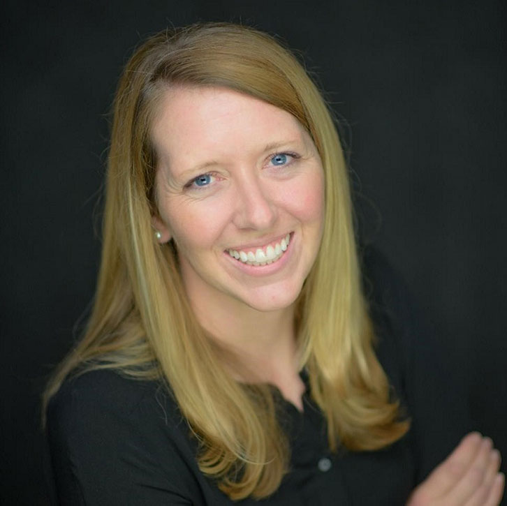 A headshot of a woman with long blonde hair, smiling in front of a dark gray background. She wears a black blouse and has her arms loosely crossed.