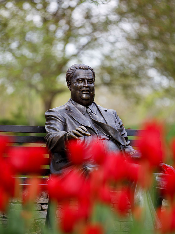 A bronze statue of Herman B Wells, smiling with one hand outstretched. The statue is surrounded by red tulips, with trees in the background.