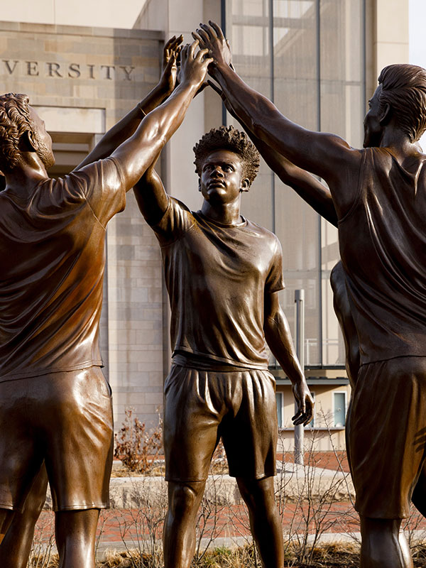 A bronze statue of four male athletes in soccer uniforms standing in a circle, reaching up to join hands. The backdrop features a university building partially visible.